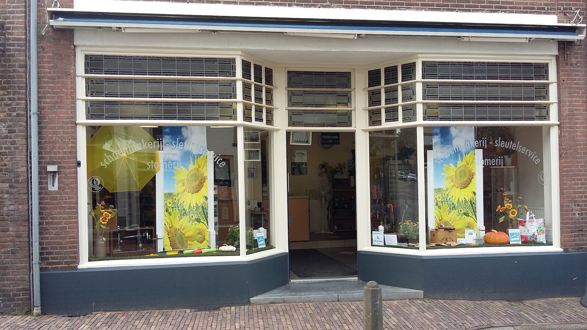 De winkelpui overdag, met zonnebloemen in de etalage en de raamletters schoenmakerij en sleutelservice